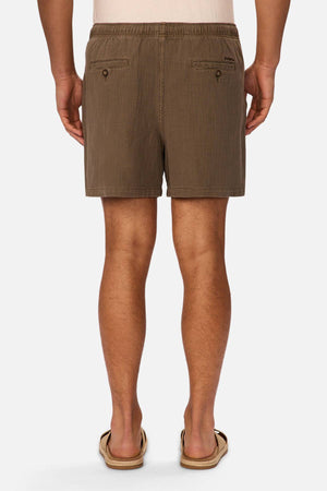 A man is seen from behind wearing Industrie The Mallorca Short, which has an elastic waist and two buttoned back pockets, paired with a light shirt and beige sandals, standing against a plain white background.