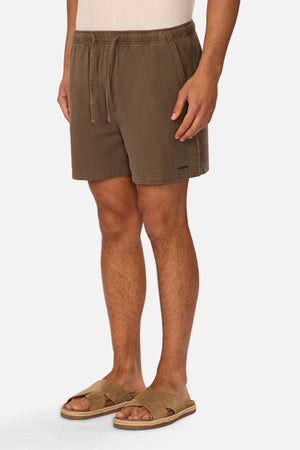 Someone stands against a white background wearing Industrie The Mallorca Short in brown, featuring an elasticated waist, paired with tan slide sandals, hands resting by their sides.