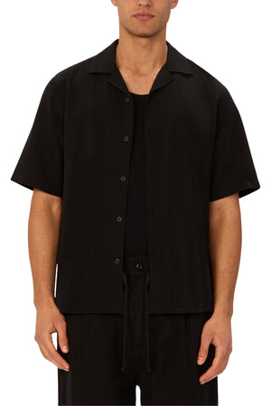 A person wears the Industrie The Marbella Short Sleeve Shirt, a relaxed fit, layered over a black top with black pants, standing against a plain white background.