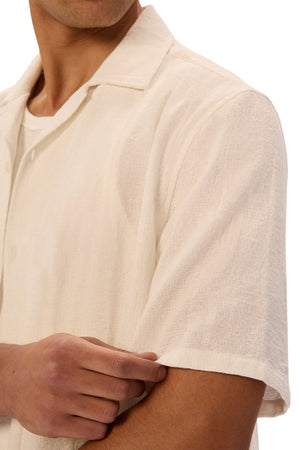 A person wears the Industrie The Marbella Short Sleeve Shirt in a relaxed fit, cream color, shown from shoulders to mid-torso, casually pulling at the sleeve; their face is partially out of frame to highlight an everyday style.