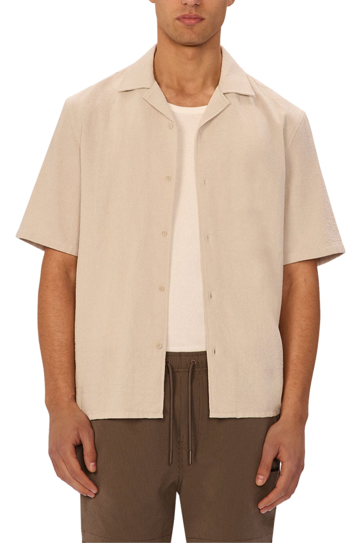 Wearing the Industrie The Marbella Short Sleeve Shirt in relaxed light beige over a white tee with brown drawstring shorts, a person stands against a plain background.