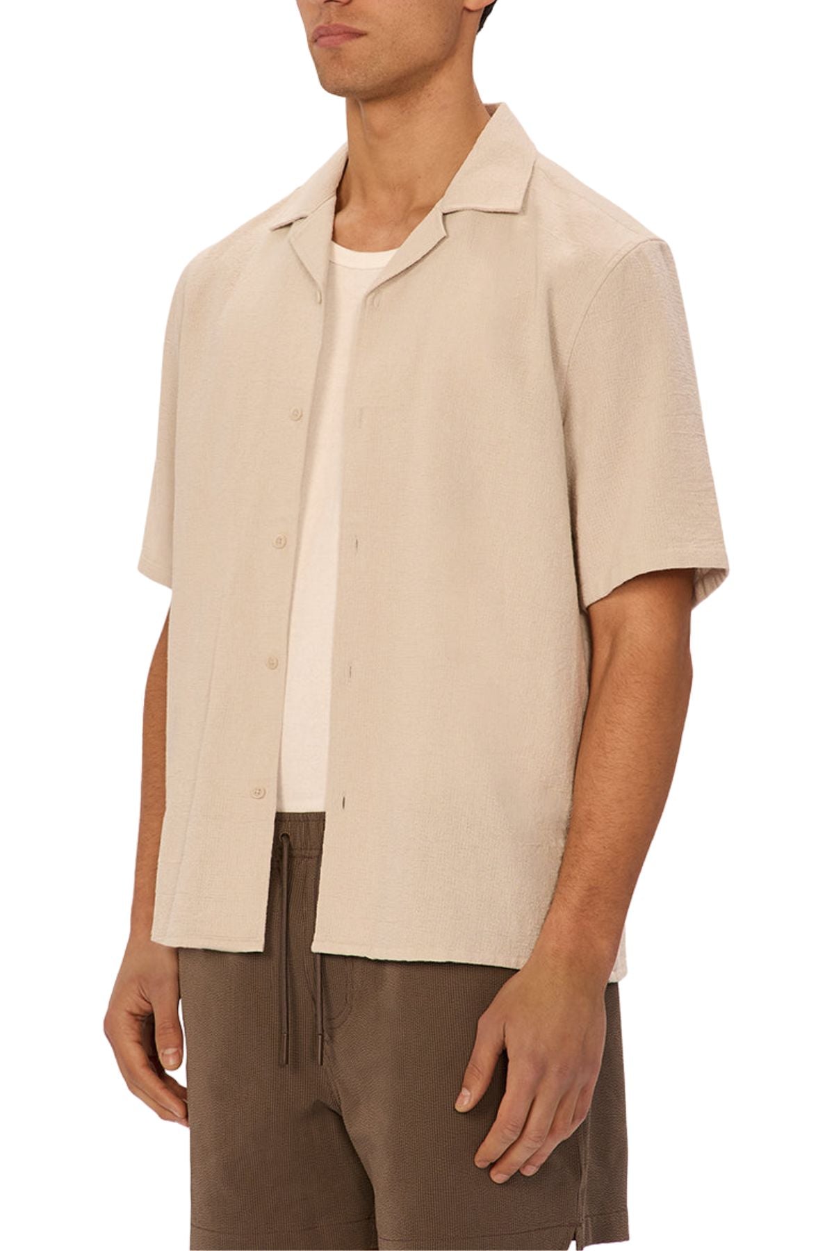 A man wears the Industrie The Marbella Short Sleeve Shirt, a relaxed-fit beige button-up in lightweight textured fabric, layered over a white T-shirt and brown drawstring shorts, standing against a plain white background.