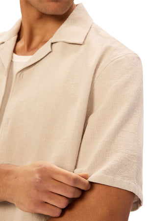 A man wearing the Industrie The Marbella Short Sleeve Shirt in light beige gently pulls at the sleeve. His face is partially out of frame, set against a plain white background.
