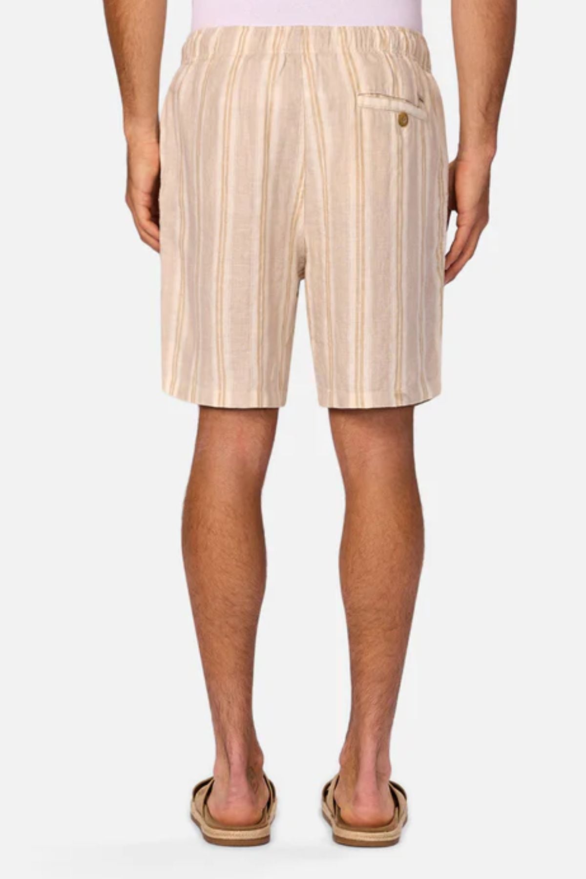 A person is shown from the back wearing Industrie The Matias Short in beige and white stripes with sandals and a light pink shirt. The breathable linen fabric and elastic waistband make these shorts ideal for everyday comfort.