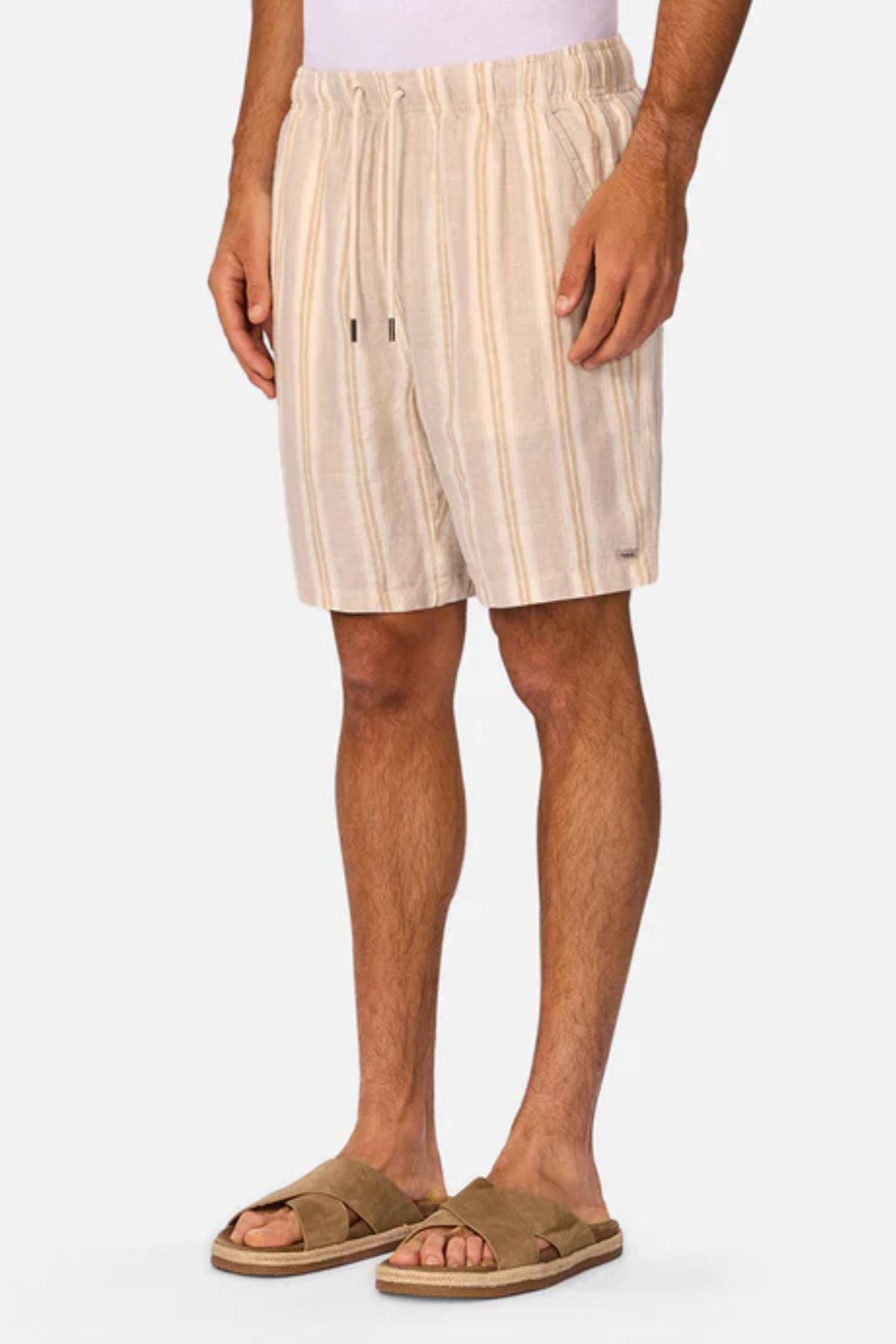 A man wears Industrie The Matias Short—beige and white striped linen shorts—in breathable fabric with tan slide sandals, standing against a light gray background. Only his body from mid-torso to feet is shown, highlighting casual style.