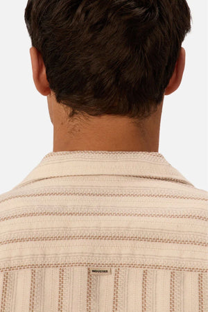 A person with short brown hair is seen from behind wearing the Industrie The Miramar Short Sleeve Shirt, a cream-colored top with light brown stripes and a small “INDUSTRIE” label on the back.