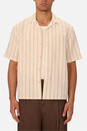 A man stands against a plain background wearing the Industrie The Miramar Short Sleeve Shirt with vertical stripes over a white T-shirt, paired with brown pants.