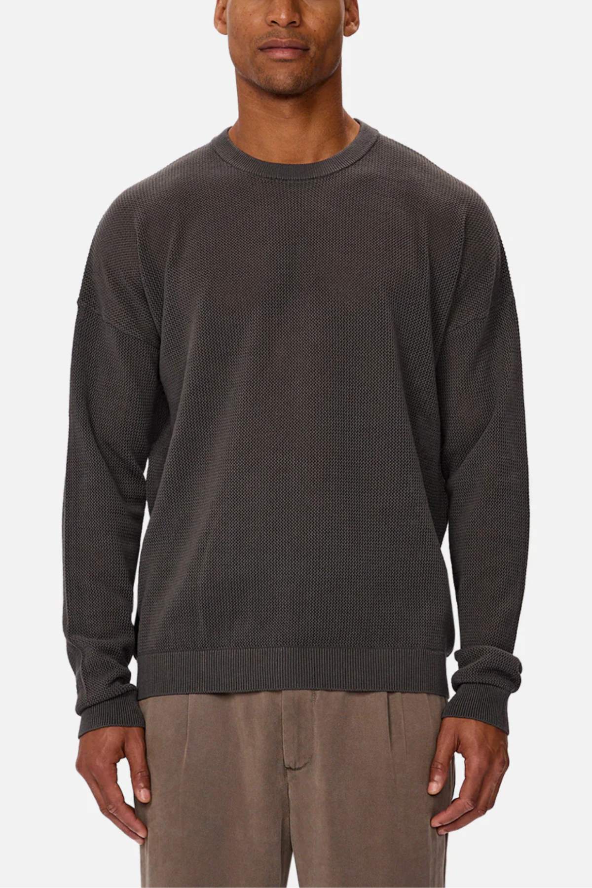 A man stands facing forward against a plain white background, wearing the Industrie The Nanterre Crew Neck Knit in dark gray with long sleeves and light brown pants.