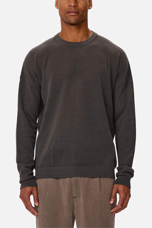 A man stands facing forward against a plain white background, wearing the Industrie The Nanterre Crew Neck Knit in dark gray with long sleeves and light brown pants.