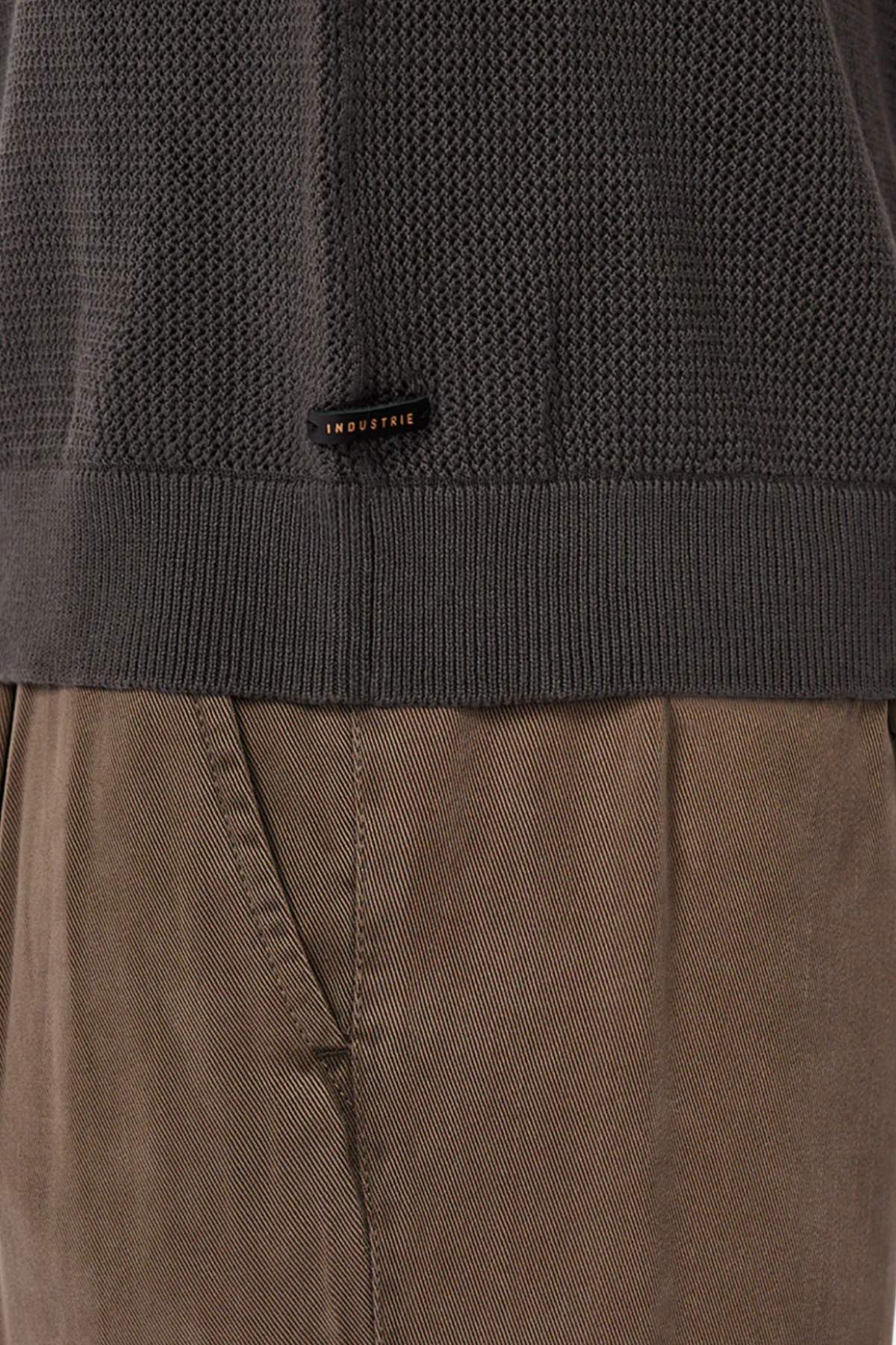 Close-up of a person wearing brown textured pants and the Industrie The Nanterre Crew Neck Knit in dark gray, featuring a small "INDUSTRIE" label near the hem. Only the lower sweater and upper pants are visible.
