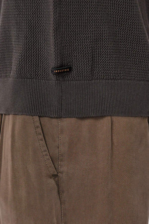 Close-up of a person wearing brown textured pants and the Industrie The Nanterre Crew Neck Knit in dark gray, featuring a small "INDUSTRIE" label near the hem. Only the lower sweater and upper pants are visible.
