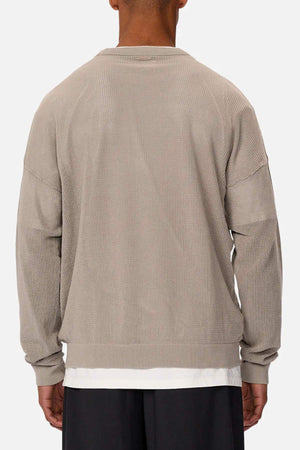 Someone is wearing Industrie The Nanterre Crew Neck Knit, showcasing an oversized fit and textured beige fabric with dropped shoulders. The long sleeves reveal a white shirt underneath, paired with dark pants against a plain backdrop for a stylish look.