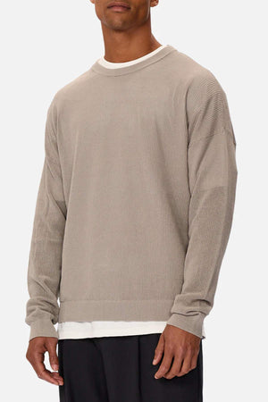 A person dons the Industrie The Nanterre Crew Neck Knit, a beige, oversized 100% cotton sweater with long sleeves over a white shirt. It's paired with black pants against a plain white background.