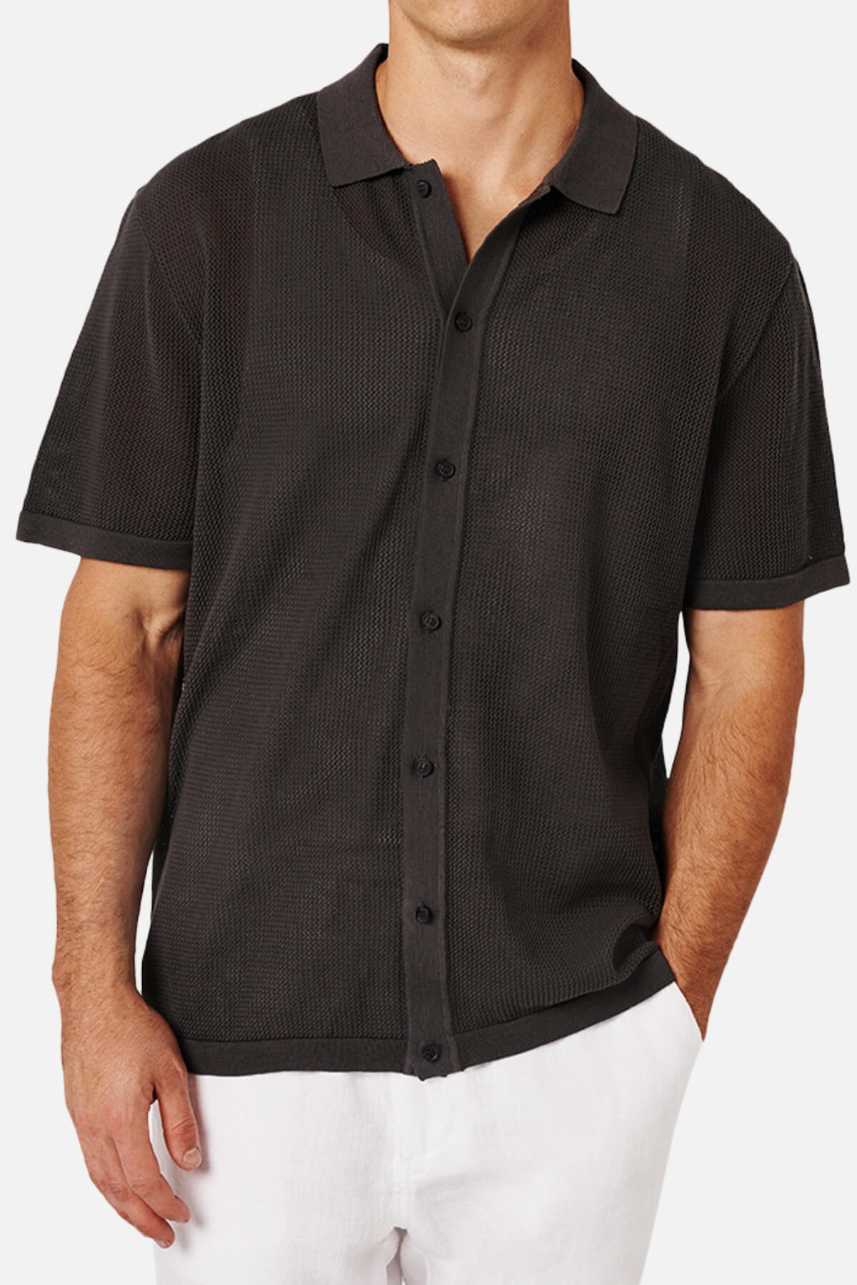 A man wears the Industrie The Nanterre Short Sleeve Shirt, a black button-up with a camp collar and textured pattern, paired with white pants. His left hand in his pocket highlights an effortlessly casual look.