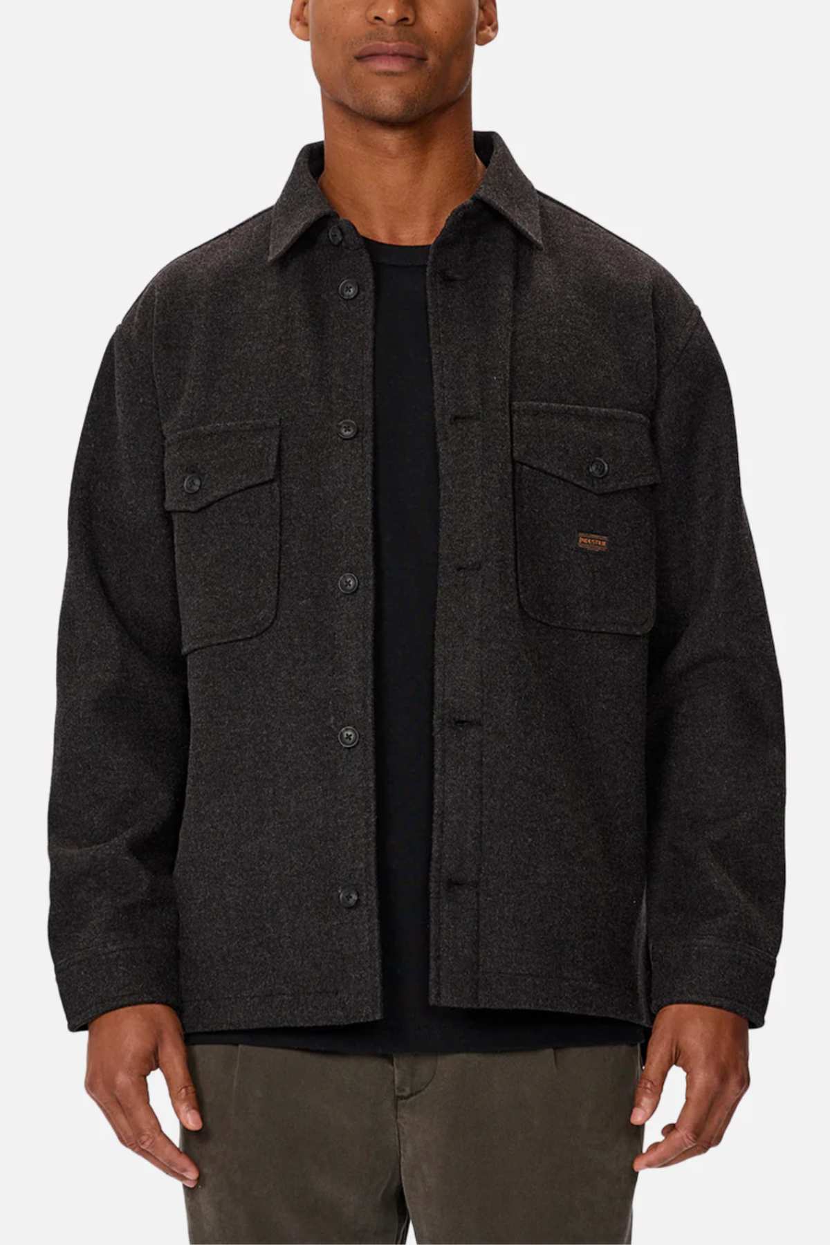 The person is dressed in an Industrie The New Coleman Jacket with a relaxed fit and classic collar, featuring two chest flap pockets. They're wearing a black shirt and olive green pants, set against a plain white backdrop that highlights the outfit's elements.