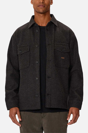 The person is dressed in an Industrie The New Coleman Jacket with a relaxed fit and classic collar, featuring two chest flap pockets. They're wearing a black shirt and olive green pants, set against a plain white backdrop that highlights the outfit's elements.