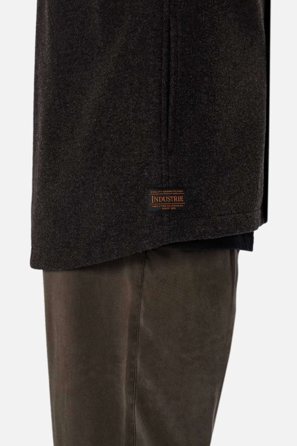 Close-up of a person wearing the Industrie The New Coleman Jacket in dark gray, showing a classic collar with the "INDUSTRIE" label near the hem, paired with olive green pants. The relaxed fit of the jacket is highlighted against a plain white background.