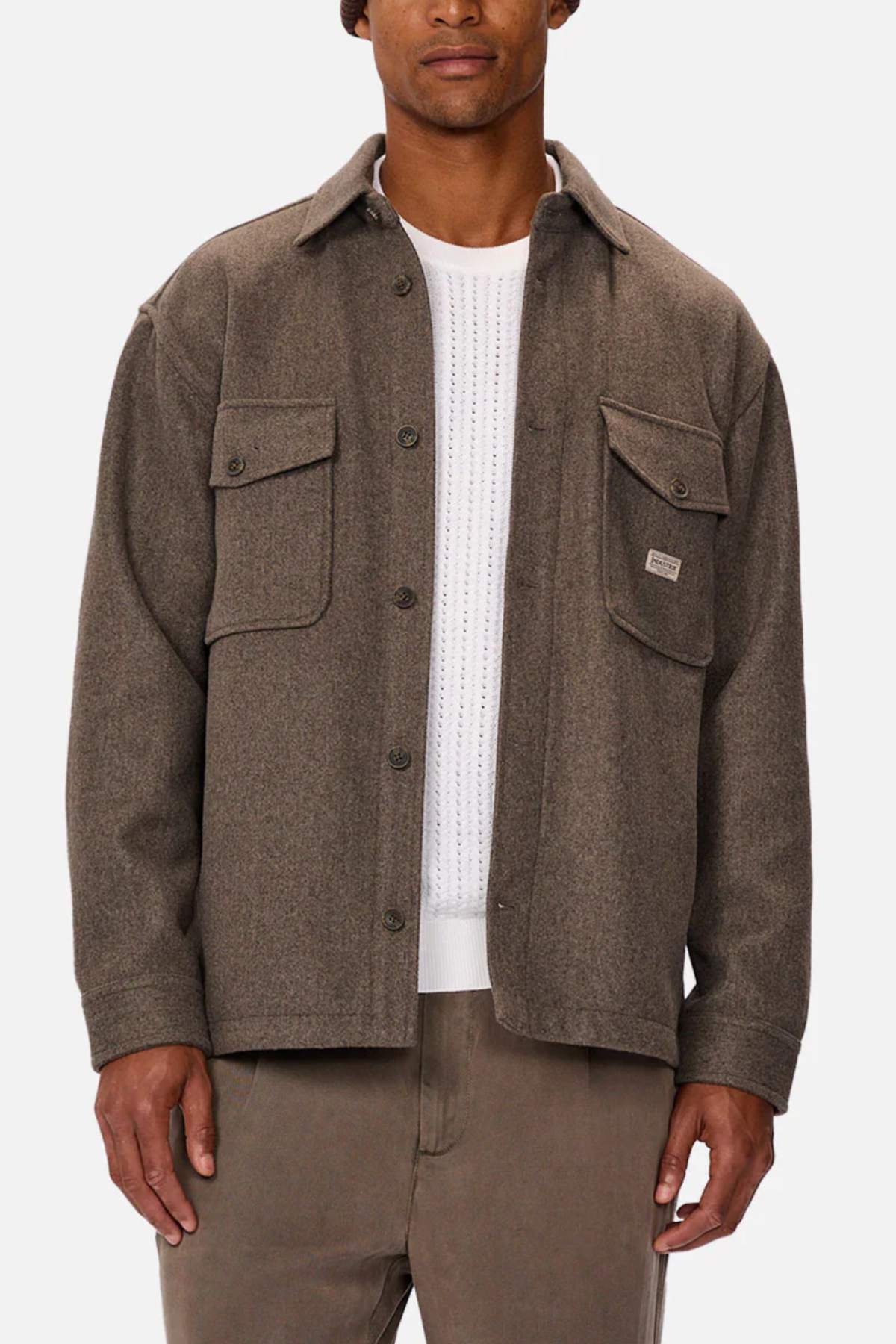 A man wears the Industrie The New Coleman Jacket, a relaxed-fit brown wool button-up with classic collar and two chest pockets, layered over a white knit sweater and brown pants, standing against a plain background.