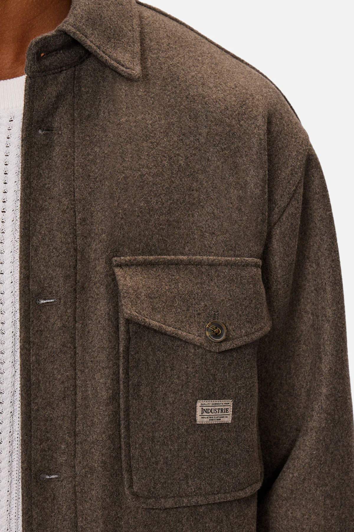 A close-up of a person wearing the Industrie The New Coleman Jacket—a dark brown wool shirt jacket with a classic collar, large buttoned chest pocket, and INDUSTRIE label—layered over a white textured sweater for a relaxed fit.