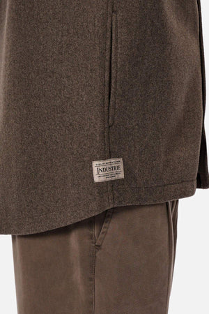 Close-up of a person wearing the Industrie The New Coleman Jacket with brown pants. The relaxed fit jacket features a classic collar and a rectangular "INDUSTRIE" label near the hem. Background is plain white.