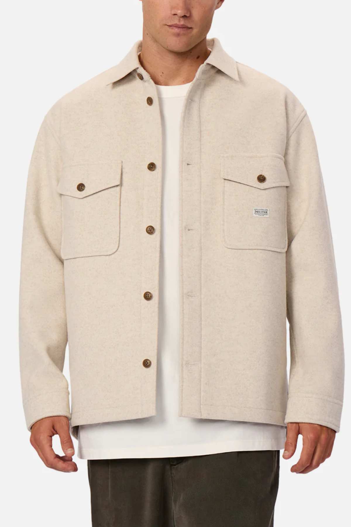 A man stands against a white background wearing a relaxed fit Industrie The New Coleman Jacket in light beige with chest pockets, layered over a white t-shirt and paired with dark pants.