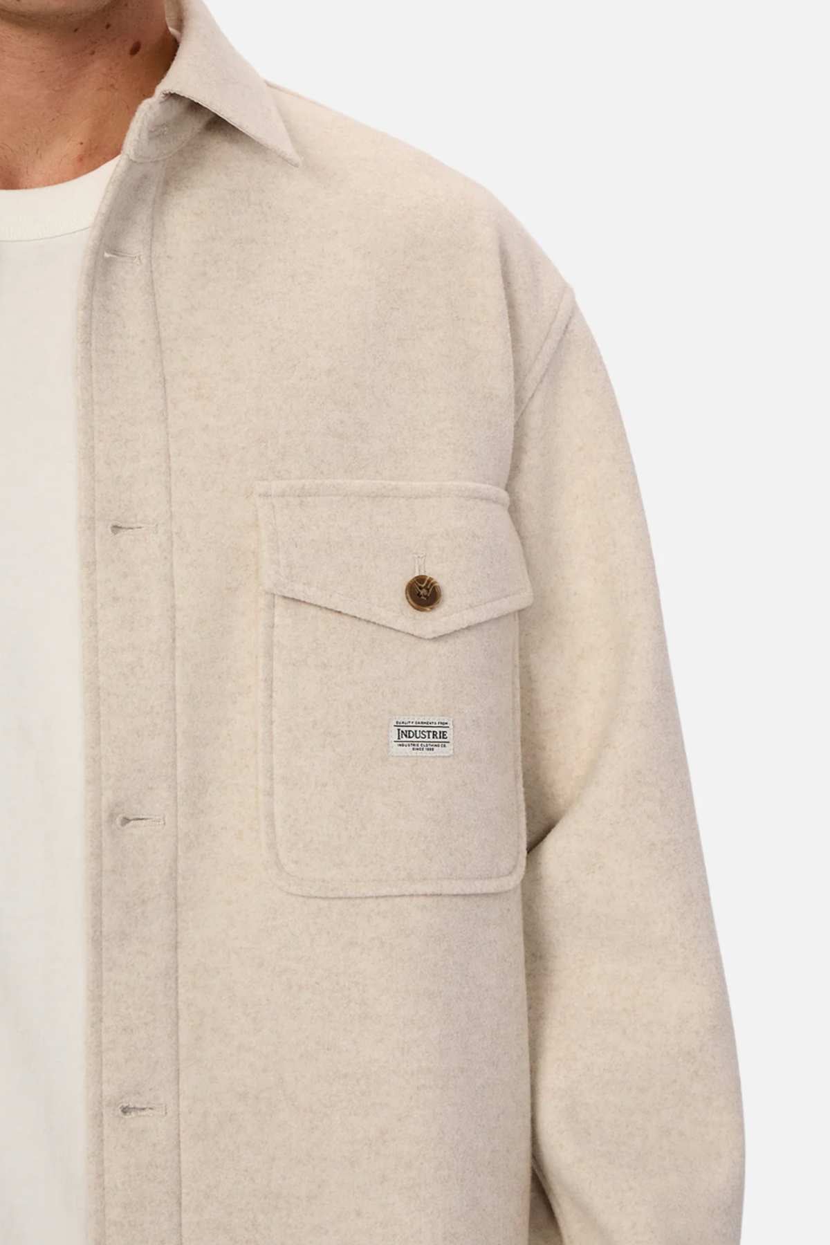 A close-up of a person wearing the Industrie The New Coleman Jacket, a cream-colored, relaxed fit button-up with a chest flap pocket, brown button, and small rectangular label. The collar and visible white T-shirt add classic cool.