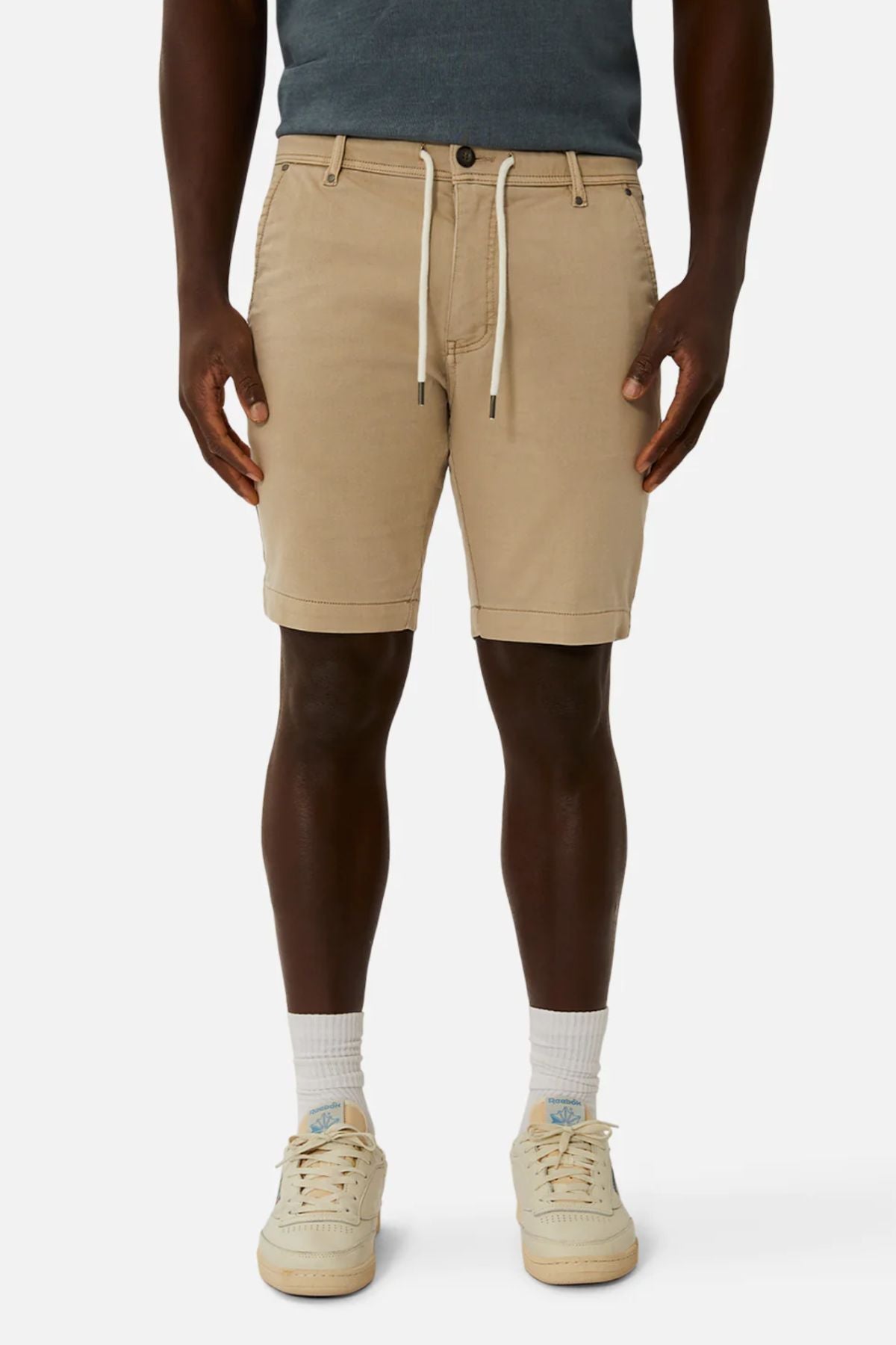 A person stands confidently against a plain white background, wearing stylish beige Industrie The New Drifter Cuba Shorts with a white drawstring, paired with a teal shirt and cream sneakers featuring blue accents.