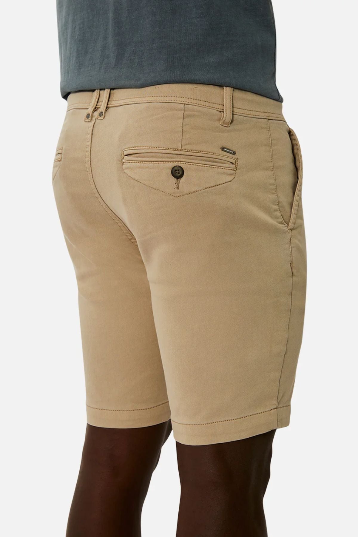 A person wearing beige Industrie The New Drifter Cuba Short, featuring side pockets and buttoned back pockets, paired with a gray top, with the lower back and legs partially visible.