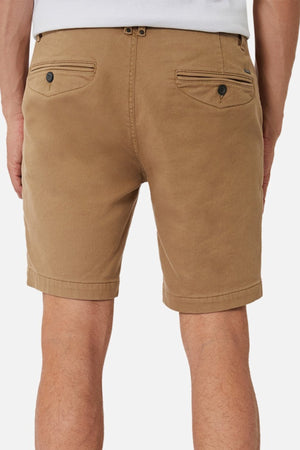 A person wears tan Industrie The New Drifter Cuba Short, featuring a casual stylish design with two buttoned back pockets and belt loops, while standing against a plain white background.