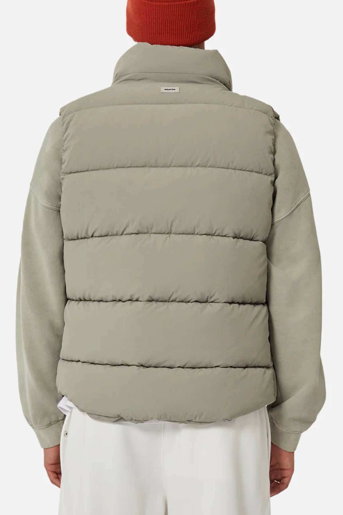 Someone wearing the Industrie The New Hanam Puffer Vest in light gray over a gray sweatshirt, paired with white pants and an orange knit beanie, is shown from the back against a plain background.