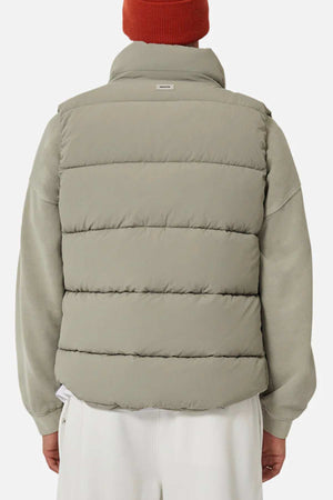 Someone wearing the Industrie The New Hanam Puffer Vest in light gray over a gray sweatshirt, paired with white pants and an orange knit beanie, is shown from the back against a plain background.