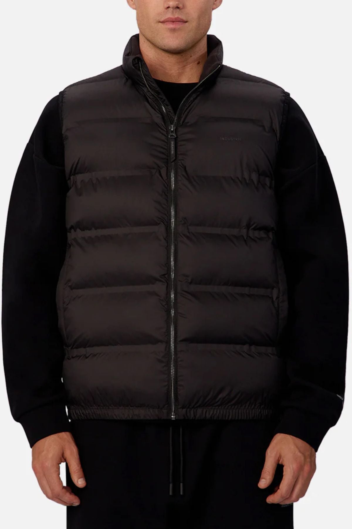 Someone stands facing forward against a plain light background, wearing the Industrie The New Kyoto Puffer Vest over a black long-sleeve shirt. The vest features horizontal quilting, heat-sealed seams, and a sleek front zipper.