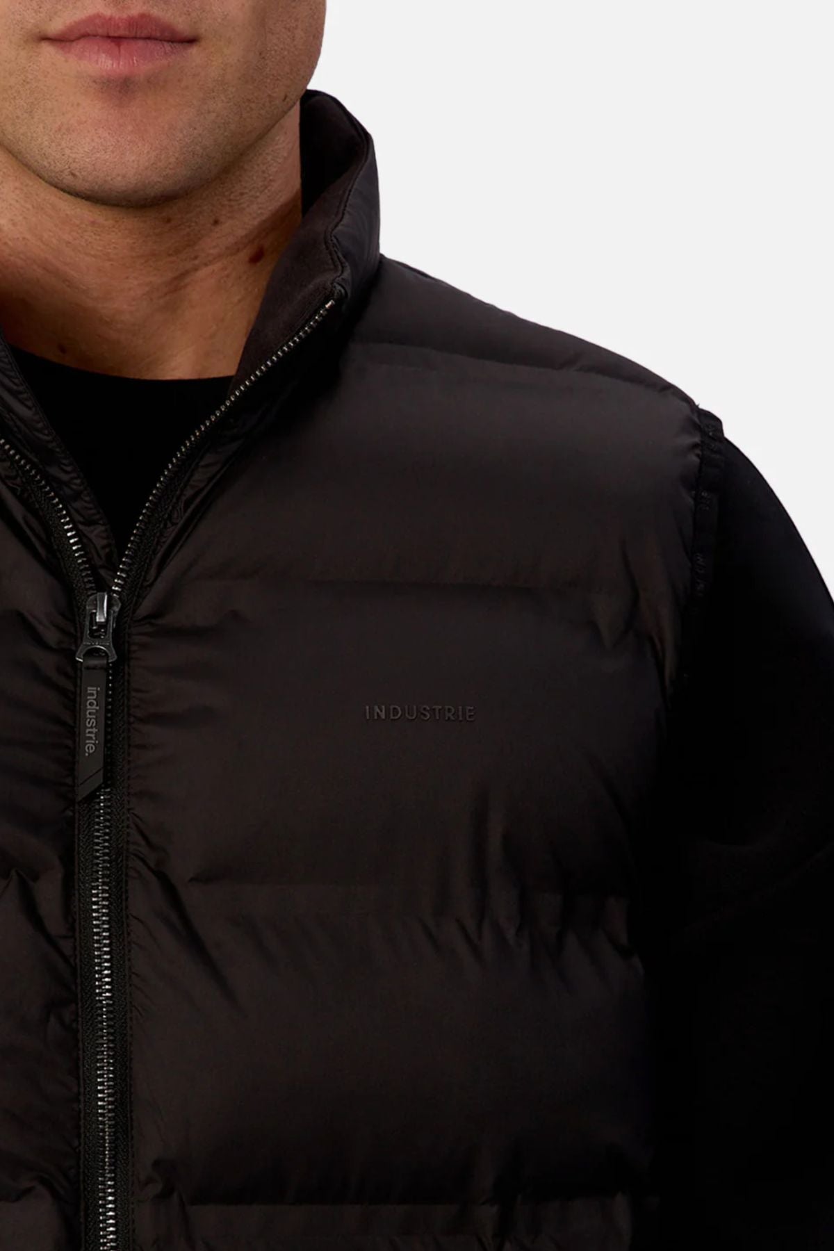 A close-up of a person wearing the Industrie The New Kyoto Puffer Vest over a black shirt. This vest has heat-sealed seams, a zipper, and "INDUSTRIE" subtly printed on the chest, set against a plain light background.