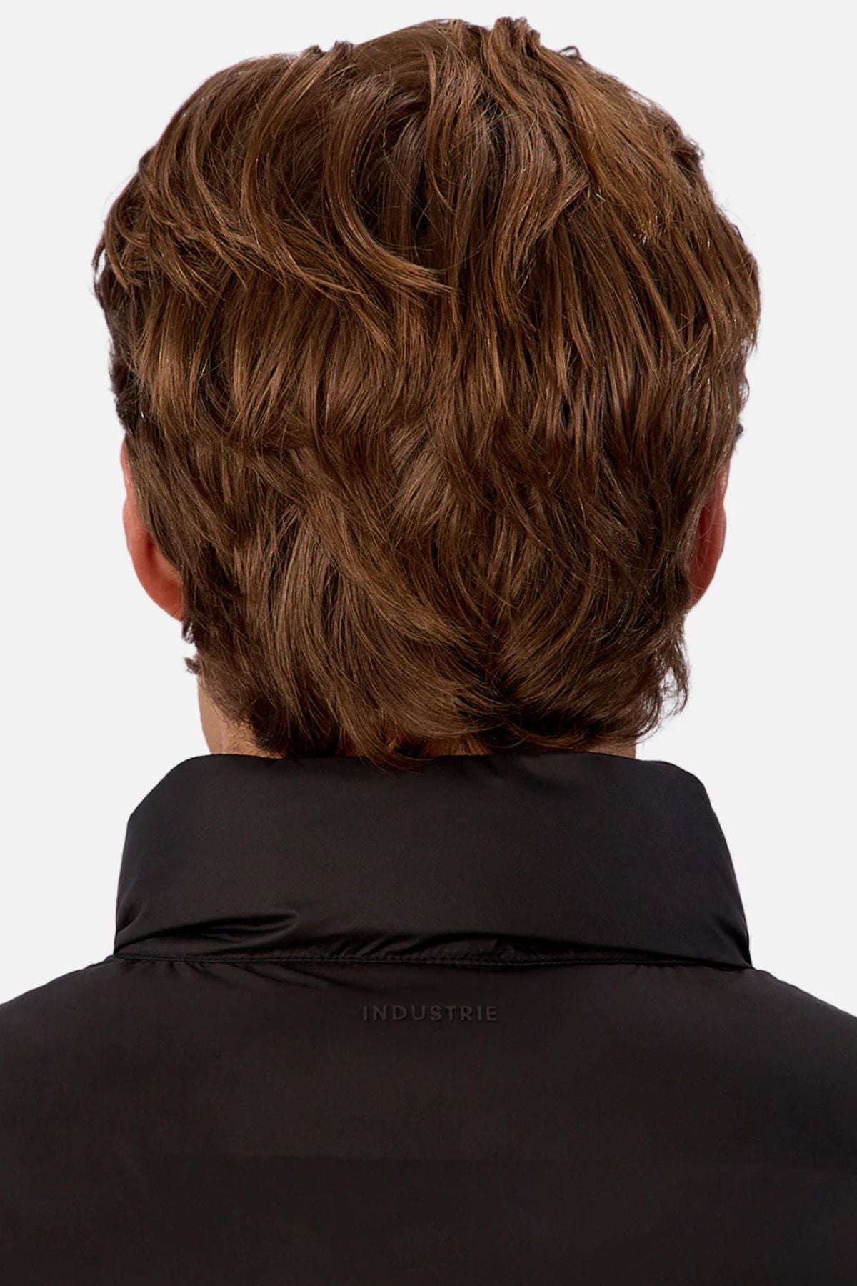 A person with wavy, light brown hair is seen from behind wearing the Industrie The New Kyoto Puffer Vest in black, which features heat-sealed seams and "INDUSTRIE" subtly printed on the collar. The background is plain white.