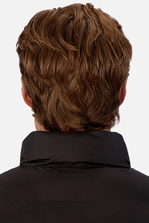 A person with wavy, light brown hair is seen from behind wearing the Industrie The New Kyoto Puffer Vest in black, which features heat-sealed seams and "INDUSTRIE" subtly printed on the collar. The background is plain white.