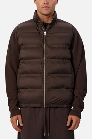 A person wears the Industrie The New Kyoto Puffer Vest in dark brown, featuring a featherlight design, layered over matching long sleeves and pants, standing against a plain light background with arms relaxedβperfect for cooler weather.