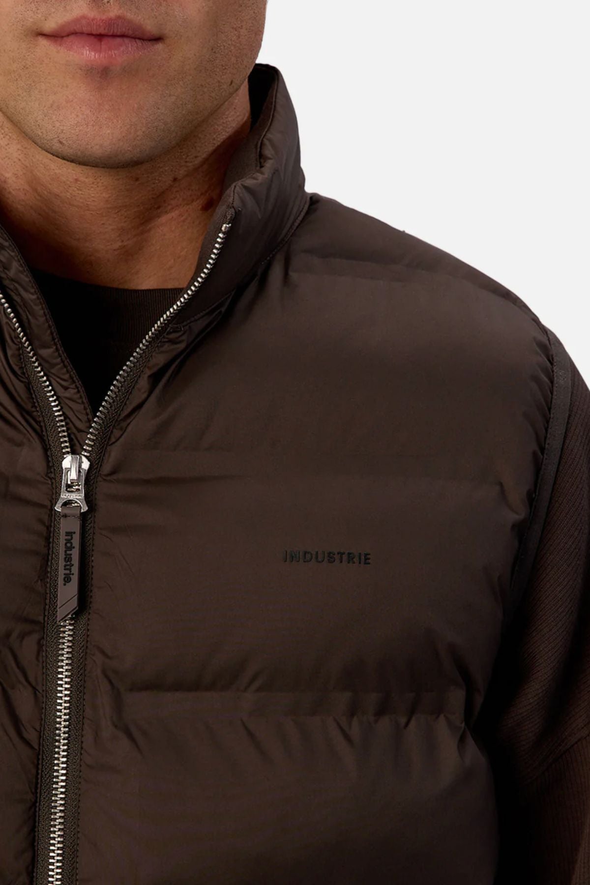 Close-up of a person wearing the Industrie The New Kyoto Puffer Vest, featuring a featherlight design, "INDUSTRIE" chest print, visible zipper, and cozy buildβideal for cooler weather against a plain, light background.
