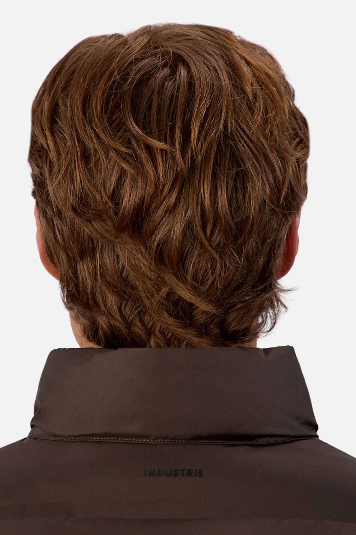The image shows the back view of a person with medium-length, wavy brown hair wearing the Industrie The New Kyoto Puffer Vest in a featherlight dark design with "INDUSTRIE" on the collar. Perfect for cooler weather, set against a plain white background.