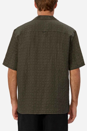 A man is seen from behind wearing the Industrie The New Ortigia Short Sleeve Shirt, which is textured in green and includes a subtle jacquard pattern with a camp collar. He pairs it with dark pants against a simple white backdrop.
