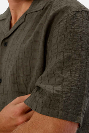 Close-up of a person wearing the Industrie The New Ortigia Short Sleeve Shirt, a textured olive green short-sleeve shirt featuring a checkered jacquard pattern. The person's arms are crossed, and the shirt showcases black buttons and a camp collar. The fabric appears lightweight and casual.
