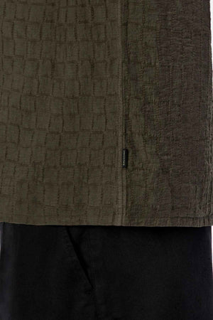 Close-up of a textured, dark green garment with a jacquard pattern, reminiscent of the Industrie The New Ortigia Short Sleeve Shirt. The camp collar design layers over black fabric, and a small tag is visible on the seam.