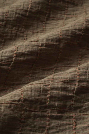 A close-up of the textured fabric with a jacquard pattern showcases a grid-like design, highlighting brown stitching on a deeper brown background. The fabric looks soft and slightly crinkled, similar to the luxurious texture found in an Industrie The New Ortigia Short Sleeve Shirt.