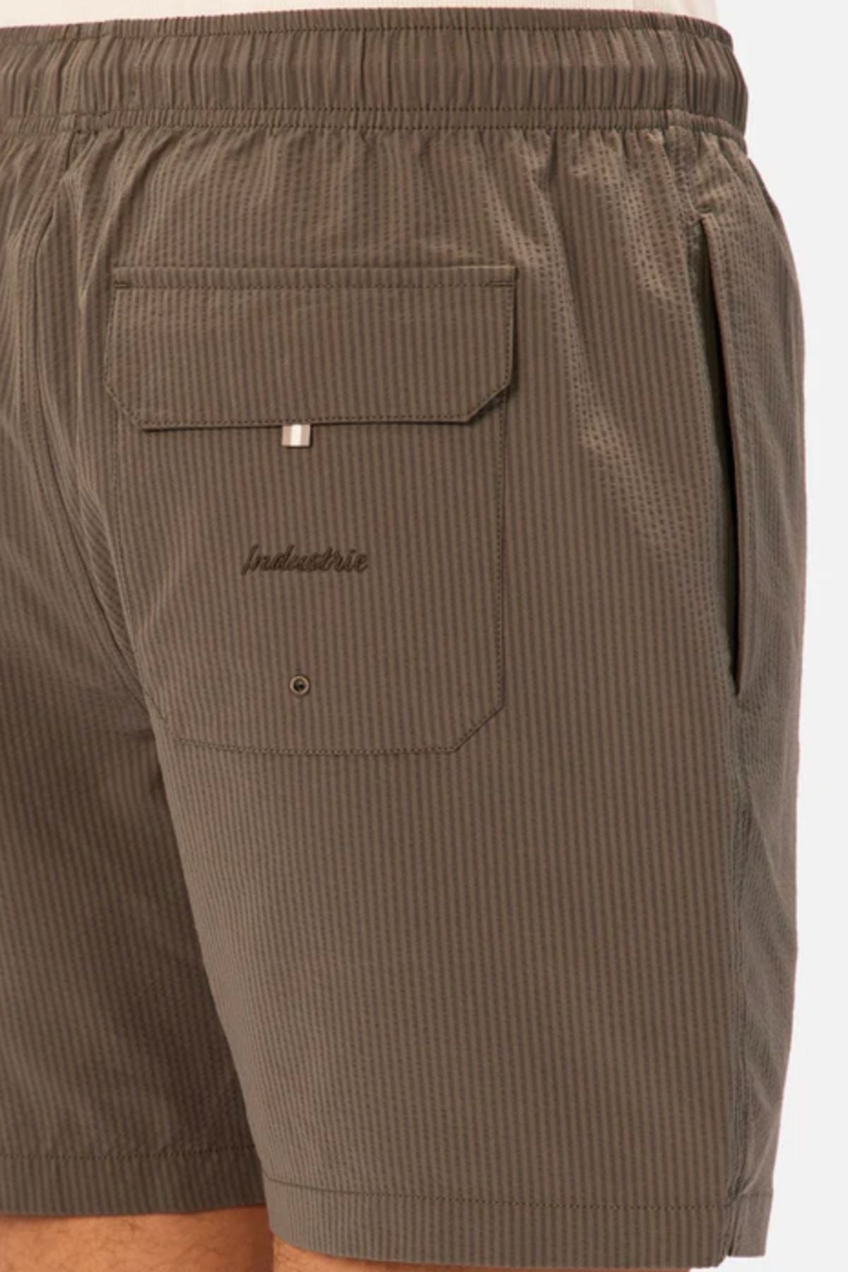 Close-up of a person wearing Industrie The New Seersucker Swim Short in brown seersucker with an elastic waistband, side pocket, and a large flap-and-button back pocket embroidered with "Industrie.