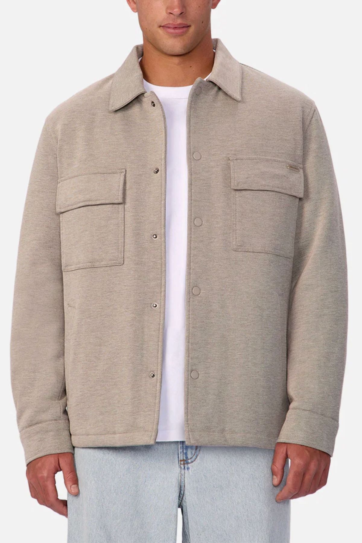 A man wears the Industrie The New Westport Jacketโa light beige casual jacket with two large chest pocketsโover a white t-shirt and light blue jeans, standing against a plain background.