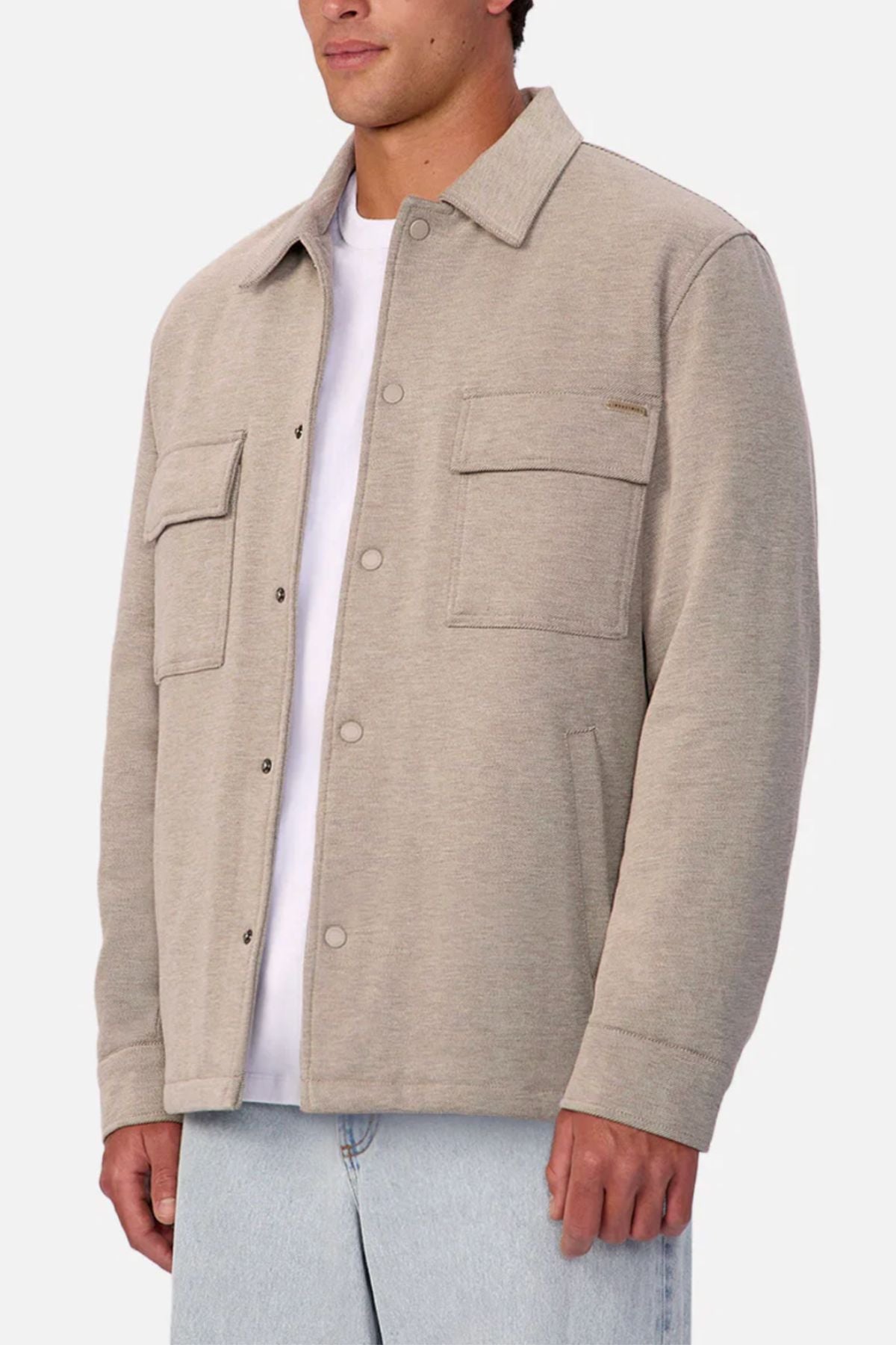 A person wears the Industrie The New Westport Jacket, a light beige casual jacket with two chest pockets, over a white T-shirt and light blue jeans, standing against a plain light background.