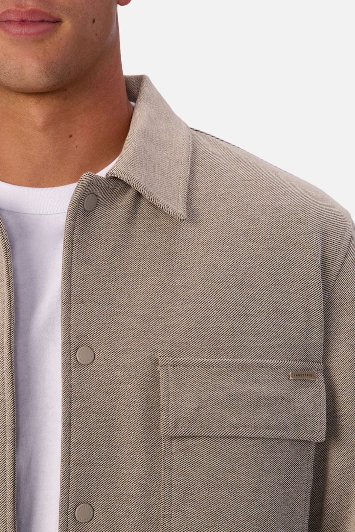 A close-up of someone wearing the Industrie The New Westport Jacketโa light beige textured casual jacket with snap buttons and a front flap pocketโover a white T-shirt, with their face partially visible.