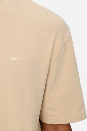 A close-up shows a person wearing the Industrie The Newport Tee in beige, featuring an oversized fit and small white "INDUSTRIE" text on the chest. Crafted from 100% cotton, the T-shirt is set against a plain, light background.
