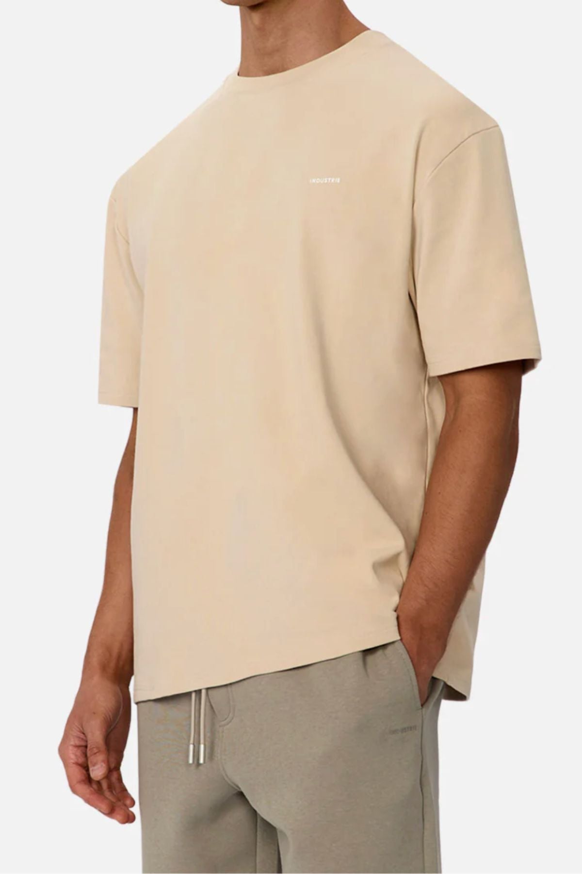 Wearing Industrie The Newport Tee in loose-fitting beige 100% cotton and light grey drawstring pants, with one hand in their pocket. Photo cropped below the nose highlights the tee's oversized, casual style.