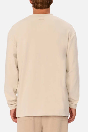 A person with short hair wearing a cream-colored Industrie The Newport Long Sleeve Tee and beige pants is seen from the back. The shirt, featuring a subtle seam detail along the shoulders, has an oversized fit and appears to have a relaxed feel.