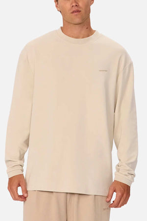 A person is wearing an oversized fit, long-sleeved light beige Industrie The Newport Long Sleeve Tee and matching light beige pants. They are standing against a plain, light background and looking forward. The shirt has a small, discrete text logo on the upper left side.